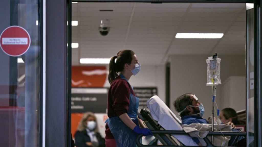 A medical professional in PPE, including gloves, an apron and a face mask as a precautionary measure against Covid-19, pushes a patient, also waering a facemask, as he lays on a bed, inside St Thomas' Hospital in north London, on April 1, 2020